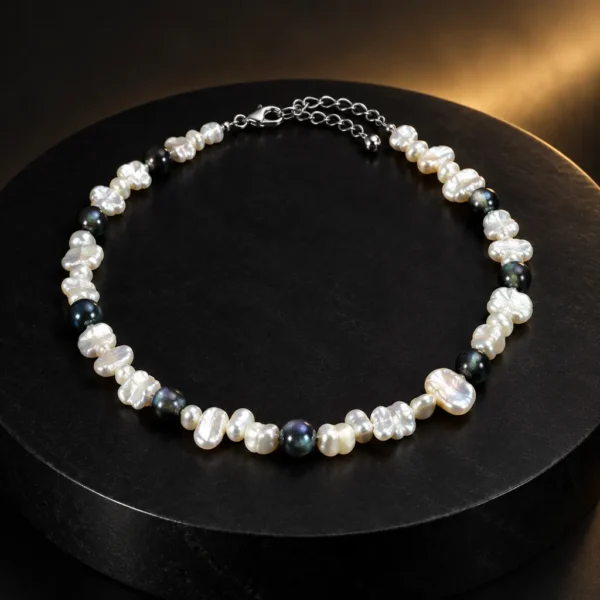 گردنبند چوکر مروارید کشی و تاهیتی نئو مینیمال Keshi & Tahitian Pearl Choker با ترکیب مروارید کشی بی شکل سفید و مروارید تاهیتی آبی مشکی و قفل خرچنگی و زنجیر رابط استیل روی استند گرد مشکی با پس زمینه تیره و نورپردازی گرم
