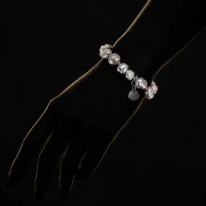 دستبند مروارید باروک و گوی های مینیمال Baroque Pearl & Silver Minimalist Bracelet با مرواریدهای باروک سفید صدفی نامنظم و گوی های نقره ای و آویز پلاک گرد نقره ای روی مچ دست زن در تاریکی با نورپردازی طلایی خطی دور دست و پس زمینه مشکی دراماتیک