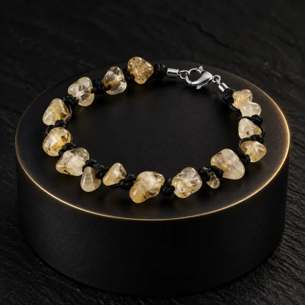دستبند راف سیترین و بند گره باف Raw Citrine Knotted Bracelet با سنگ های راف سیترین طبیعی زرد طلایی و عسلی نامنظم شفاف و بند بافت مشکی گره ای و قفل خرچنگی استیل نقره ای روی استند استوانه ای مشکی مات با لبه طلایی روی سنگ اسلیت تیره با نورپردازی لوکس
