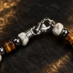 گردنبند چشم ببر و هاولیت نئو مینیمال Neo-Minimal Tiger's Eye & Howlite Choker با مهره های راف هاولیت سفید رگه دار و حدید گان متال براق و چشم ببر قهوه ای طلایی و قفل خرچنگی استیل نقره ای و حلقه فنری کلوزآپ روی سنگ اسلیت مشکی مات با نورپردازی گرم طلایی لوکس