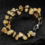 دستبند راف سیترین و بند گره باف Raw Citrine Knotted Bracelet با سنگ های راف سیترین طبیعی زرد طلایی و عسلی شفاف نامنظم و بند بافت مشکی گره ای دست ساز و قفل خرچنگی استیل نقره ای براق روی سنگ اسلیت مشکی مات با نورپردازی لوکس و پس زمینه تیره