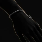 دستبند هایبرید حدید و زنجیر با قفل ملوان Gunmetal Hematite & Chain Sailor Bracelet با مهره های حدید گان متال مشکی براق و اسپیسیر های نقره ای و زنجیر استیل و قفل ملوانی حلقه ای روی مچ دست زن در تاریکی با نورپردازی طلایی خطی دور دست و پس زمینه مشکی دراماتیک هنری