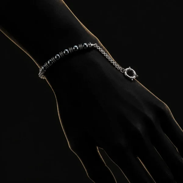 دستبند هایبرید حدید و زنجیر با قفل ملوان Gunmetal Hematite & Chain Sailor Bracelet با مهره های حدید گان متال مشکی براق و اسپیسیر های نقره ای و زنجیر استیل و قفل ملوانی حلقه ای روی مچ دست زن در تاریکی با نورپردازی طلایی خطی دور دست و پس زمینه مشکی دراماتیک هنری