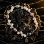 دستبند مروارید باروک و گوی های مینیمال Baroque Pearl & Silver Minimalist Bracelet با مرواریدهای باروک طبیعی سفید و صورتی بنفش صدفی و گوی های نقره ای براق و قفل خرچنگی و آویز پلاک گرد روی سنگ معدنی تورمالین سیاه براق با نورپردازی گرم طلایی و پس زمینه مخمل مشکی لوکس