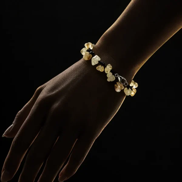 دستبند راف سیترین و بند گره باف Raw Citrine Knotted Bracelet با خوشه های سنگ راف سیترین زرد عسلی شفاف و بند بافت مشکی گره ای و قفل خرچنگی استیل روی مچ دست زن در تاریکی با نورپردازی طلایی سایه دار و پس زمینه مشکی دراماتیک