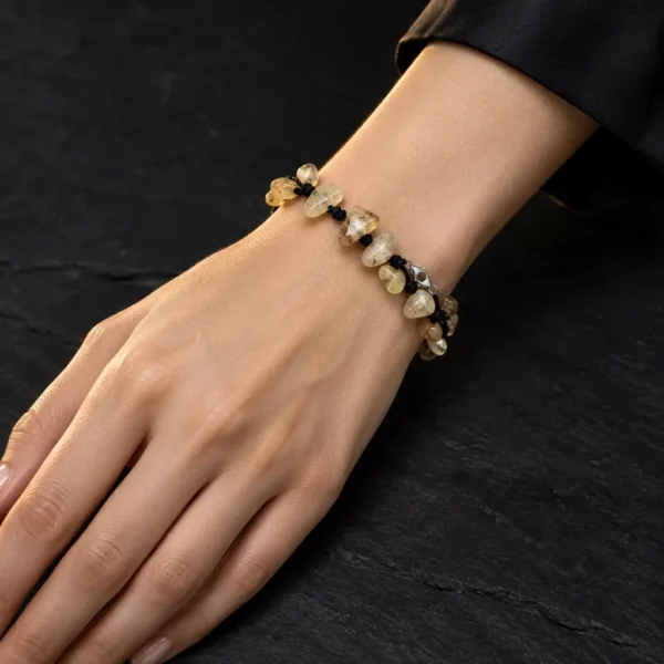 دستبند راف سیترین و بند گره باف Raw Citrine Knotted Bracelet با سنگ های راف سیترین زرد طلایی نامنظم و بند بافت مشکی گره ای و قفل خرچنگی استیل روی مچ دست زن با پوست روشن و لباس مشکی روی سنگ اسلیت مشکی مات با نورپردازی گرم طبیعی