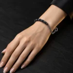 دستبند هایبرید حدید و زنجیر با قفل ملوان Gunmetal Hematite & Chain Sailor Bracelet با مهره های حدید گان متال مشکی براق و مهره های دیسکی مشکی و زنجیر استیل نقره ای و قفل ملوانی روی مچ دست زن با ناخن های نود براق و لباس مشکی روی سنگ اسلیت مشکی مات با نورپردازی لوکس
