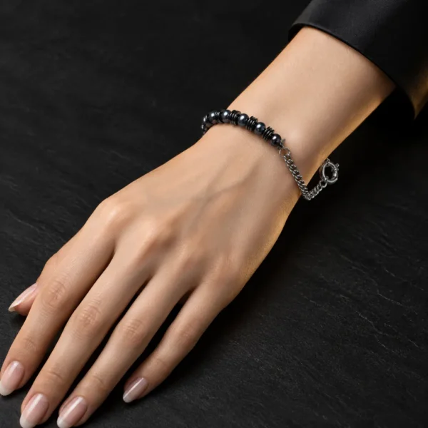 دستبند هایبرید حدید و زنجیر با قفل ملوان Gunmetal Hematite & Chain Sailor Bracelet با مهره های حدید گان متال مشکی براق و مهره های دیسکی مشکی و زنجیر استیل نقره ای و قفل ملوانی روی مچ دست زن با ناخن های نود براق و لباس مشکی روی سنگ اسلیت مشکی مات با نورپردازی لوکس